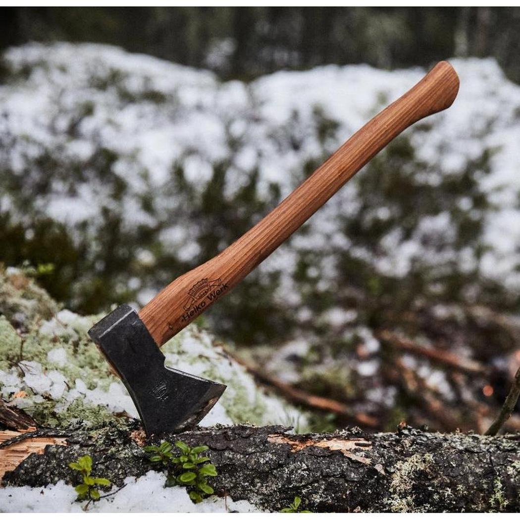 A Helko Werk Rheinlander Pack Axe with a wooden handle rests on a snow-dusted log in a forested winter setting.