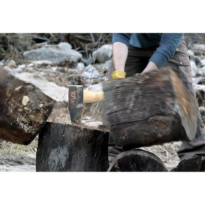 A person wearing a blue shirt and brown pants uses the Helko Werk Saxon Splitter to split a log on a tree stump in a rocky outdoor setting.