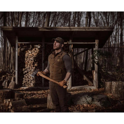 A man in a forest setting holds a Helko Werk Saxon Splitter axe, with a woodshed and stacked firewood in the background.