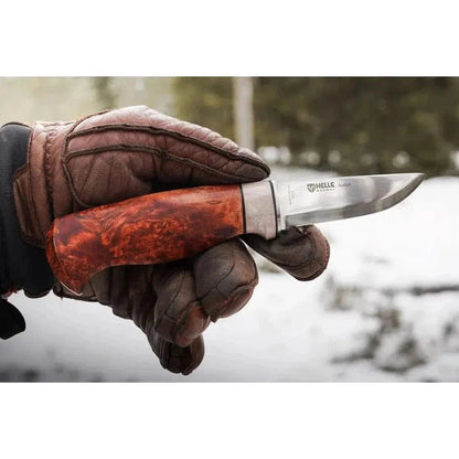 A hand in brown gloves holds the Helle Audun - 2024 LTD knife with a red handle, showing the blade and branding, against a snowy outdoor background.