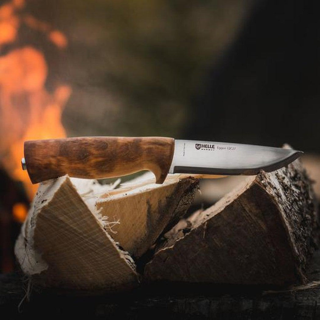 A side view of the Helle Eggen 12C27 knife with a wooden handle resting on split logs in front of a fire.