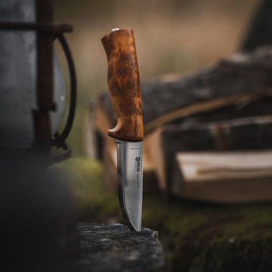 A close-up of the Helle Eggen 12C27 knife with a wooden handle, standing upright on a rock with a blurred outdoor background.