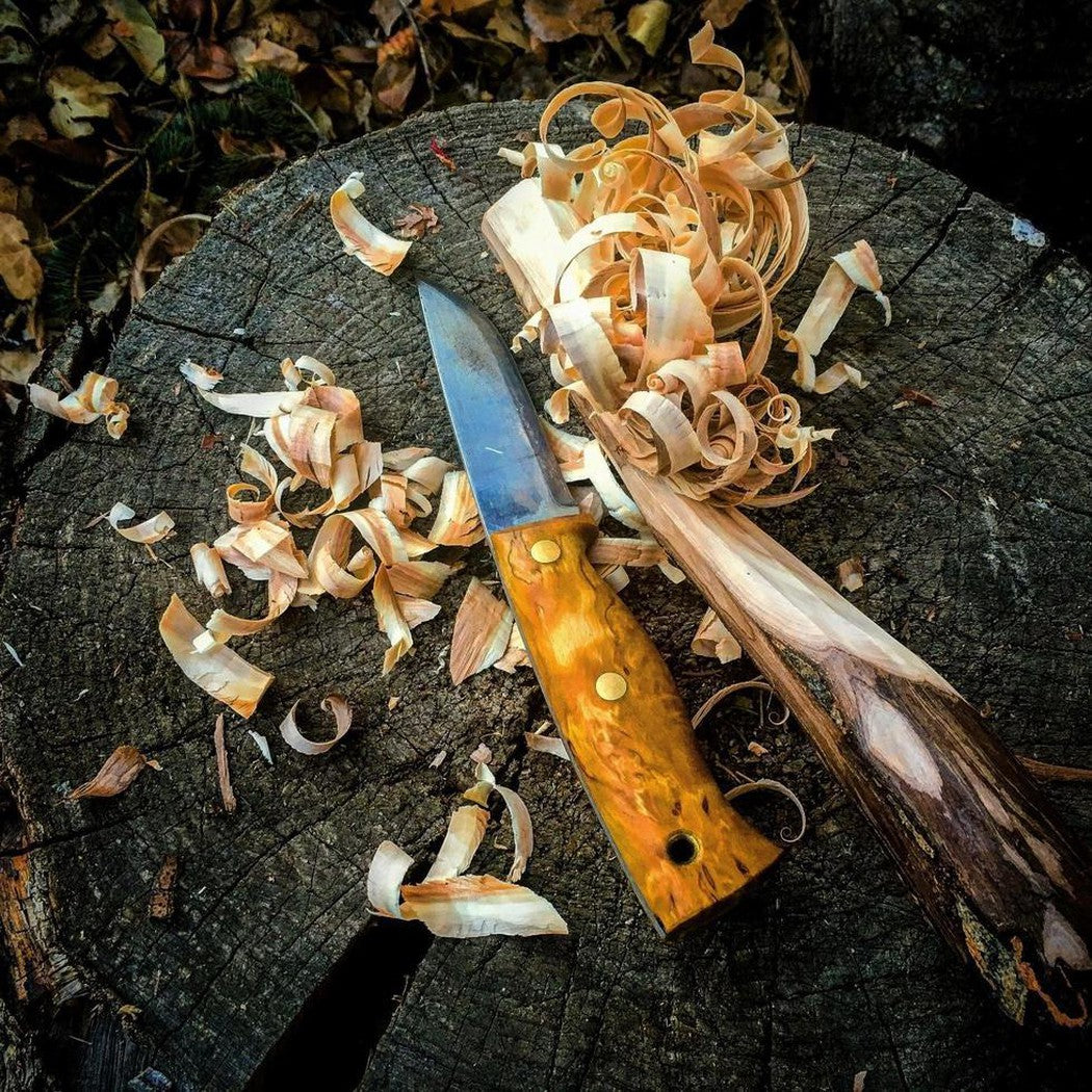 A Helle Temagami Carbon knife with a yellow handle rests on a tree stump surrounded by wood shavings.