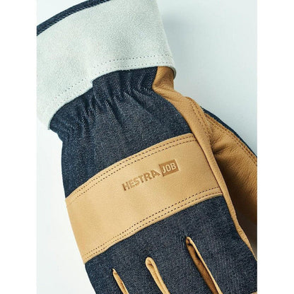 A close-up of the Hestra Job Classic Denim work glove with indigo denim and tan leather accents, showing the cuff and logo band against a light background.