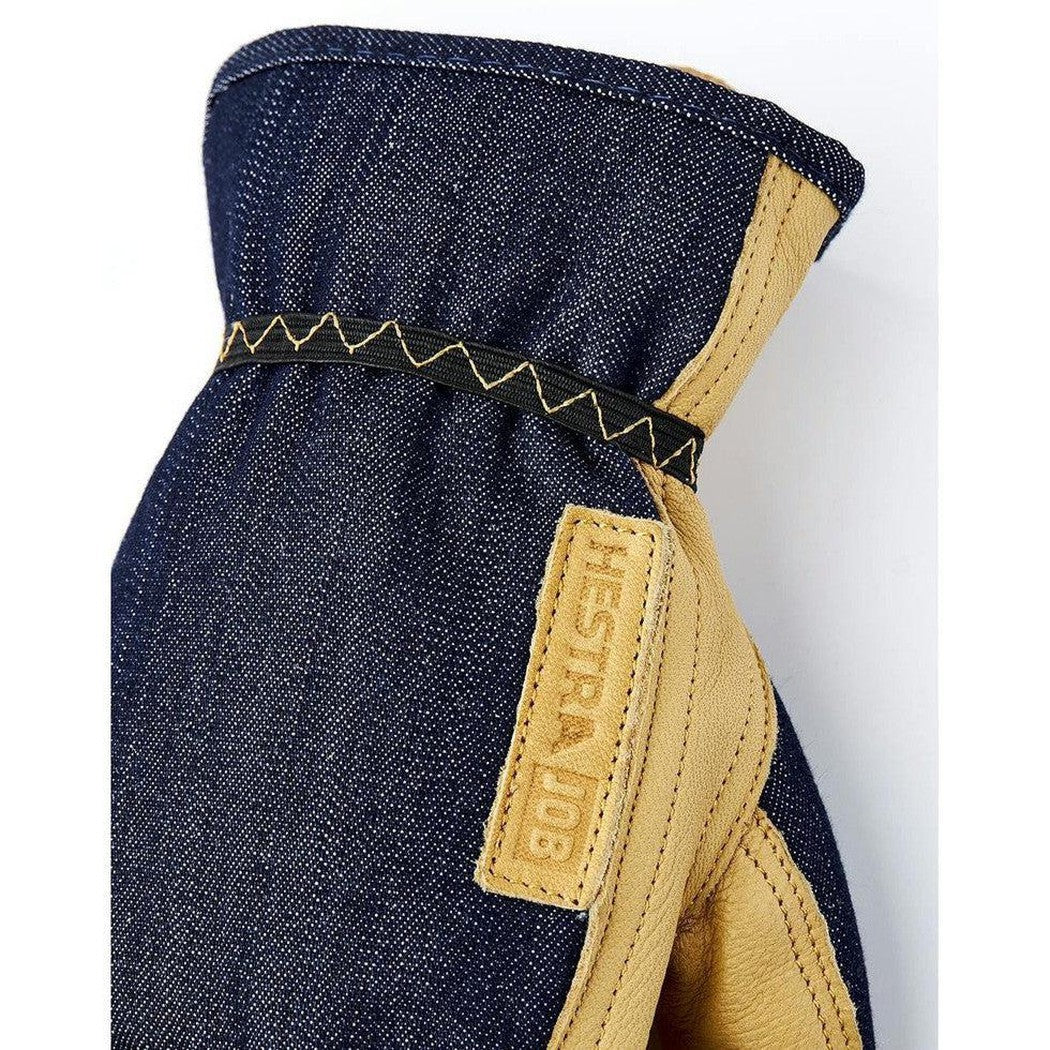 A close-up of the Hestra Job Kobolt Denim work glove in indigo denim with tan leather accents and a branded tag, photographed against a white background.