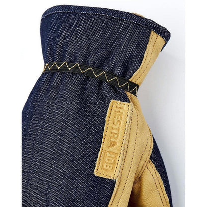 A close-up of the Hestra Job Kobolt Denim work glove in indigo denim with tan leather accents and a branded tag, photographed against a white background.