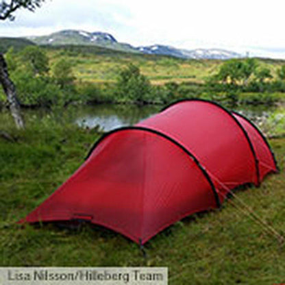 A red Hilleberg Anjan 3 GT tent sits on grass near a lake with mountains in the background.