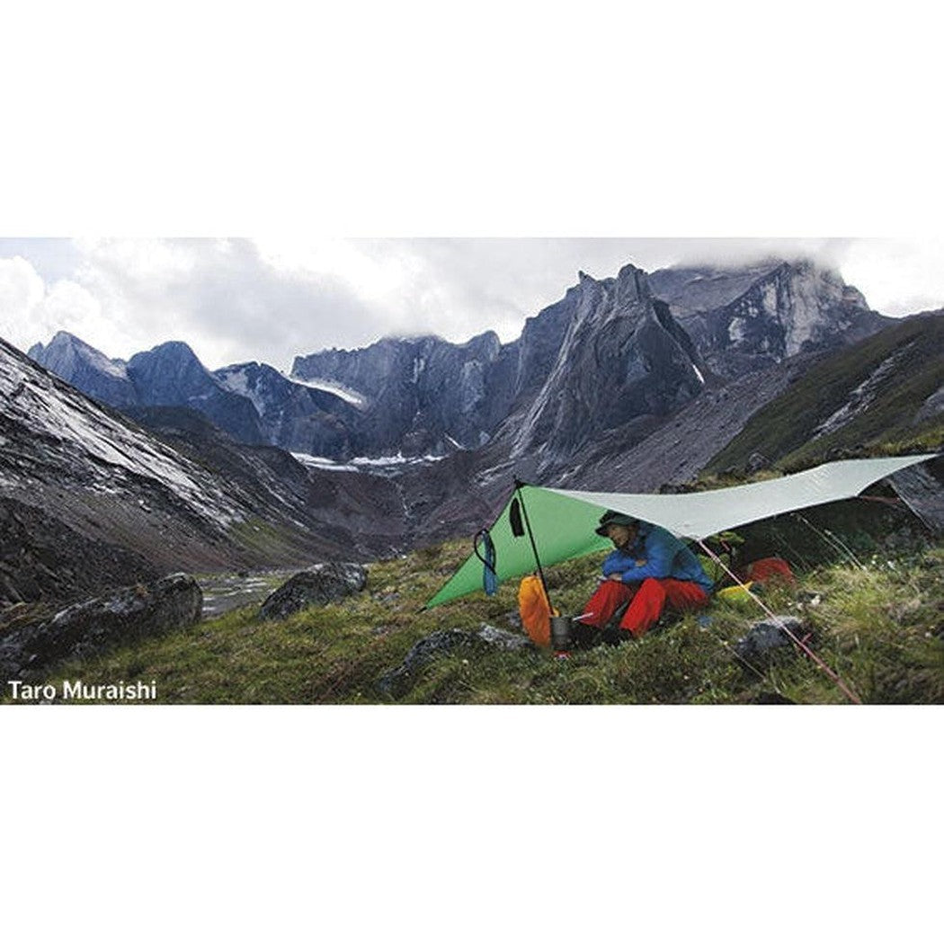 A person in blue and red sits under a green Hilleberg Tarp XP on a grassy mountain slope with rocky peaks in the background.