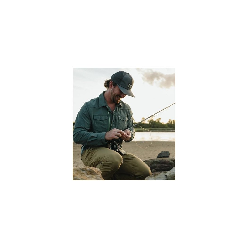 A man wearing the Faded Olive Howler Brothers Aperture Tech Strapback hat sits by a lake, adjusting his fishing gear.
