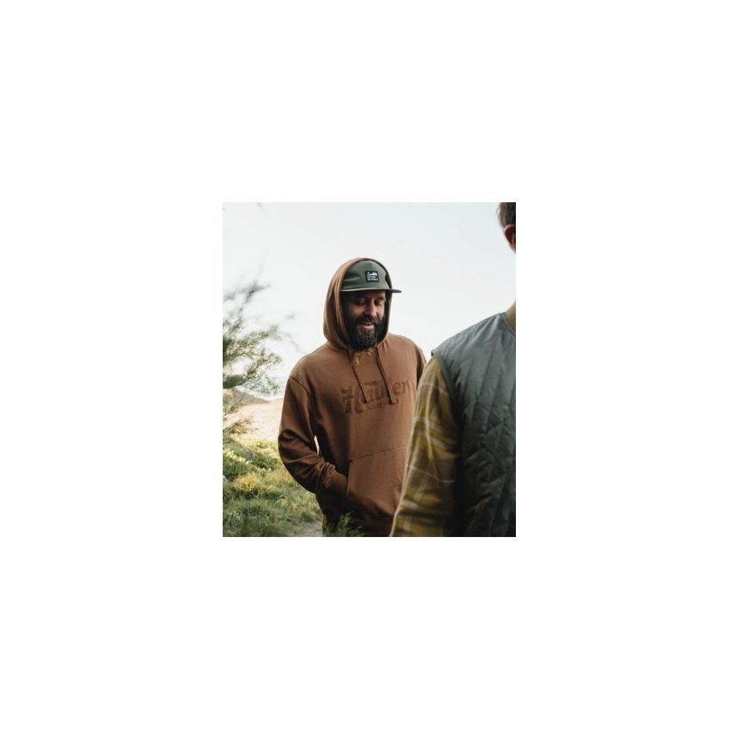 A man wears the Faded Olive Howler Brothers Aperture Tech Strapback while standing outdoors with trees in the background.