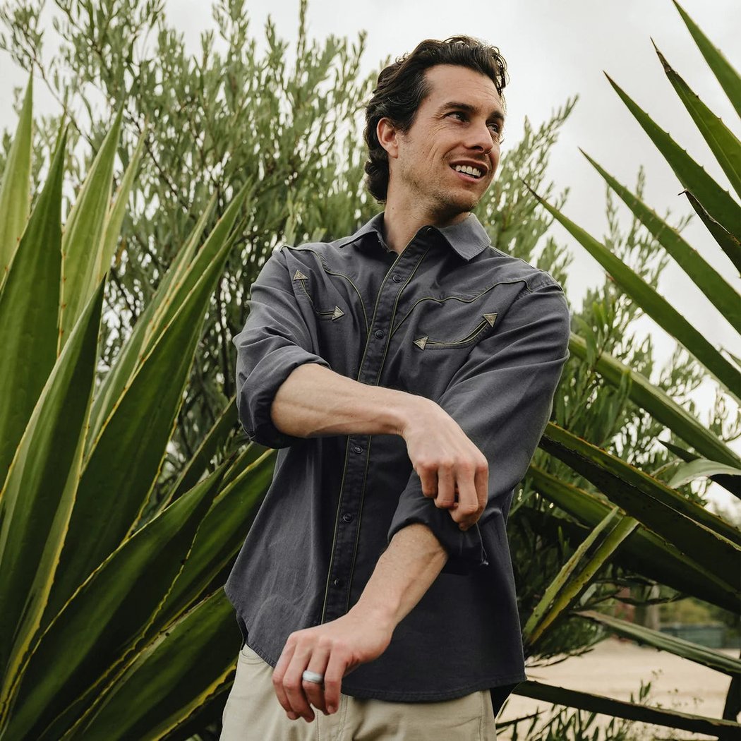 A man wears the Howler Brothers Broncbuster Longsleeve Snapshirt in MoonlessNight, rolling up his sleeve, with green foliage in the background.