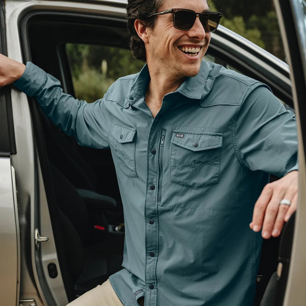 A man wearing the Howler Brothers Emerger Tech Longsleeve in Stormy Sea steps out of a car with a smile.