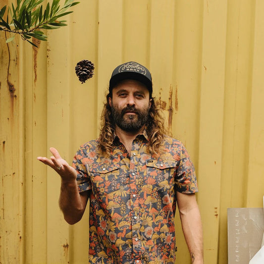A man in a Howler Lasso floral shirt and Howler Brothers Foam Dome hat tosses a pinecone in front of a yellow corrugated wall.