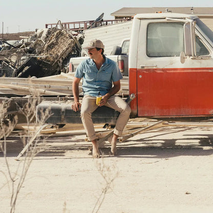A man sits barefoot on a truck bed wearing Faded Khaki Howler Brothers Frontside 5-Pocket Pants in a desert junkyard setting.
