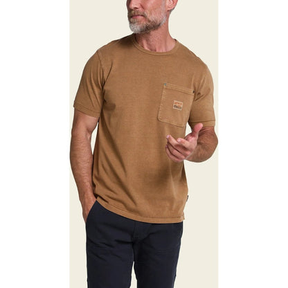 A man wearing the Howler Brothers HB Trade T - Shirt in Duck Brown stands against a light background with one hand in his pocket.