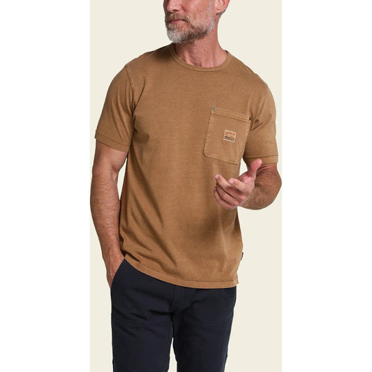 A man wearing the Howler Brothers HB Trade T - Shirt in Duck Brown stands against a light background with one hand in his pocket.
