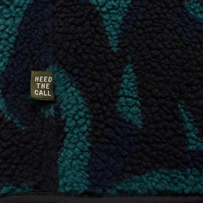 A close-up of the Howler Brothers Men's Chisos Fleece Vest in Forgotten Forest : Antique Black showing the "Heed The Call" tag against a textured pattern.
