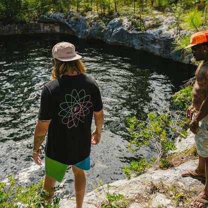 Two men stand by a rocky pool, one wearing the Howler Brothers Men's Cotton Pocket T in Black with a colorful atom graphic on the back.