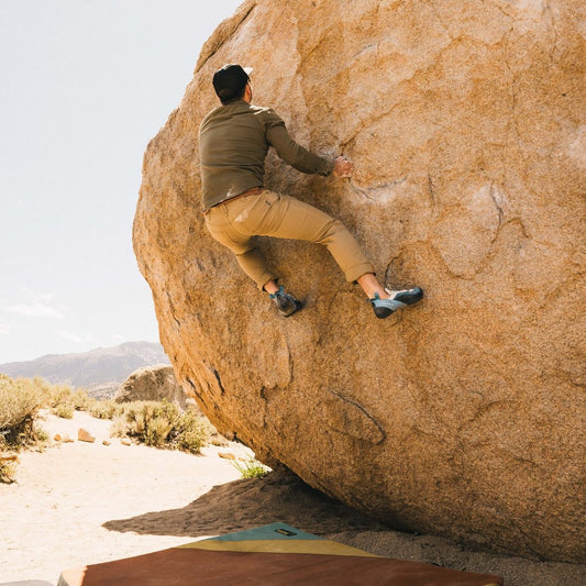 A man climbs a large rock wearing Howler Brothers Men's Frontside 5-Pocket Pants in Faded Khaki, with desert scenery in the background.