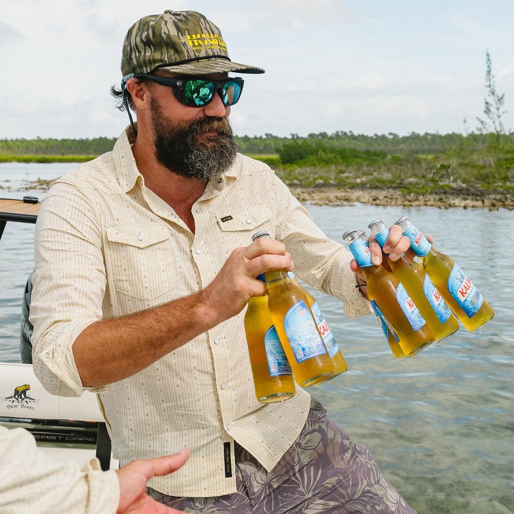 A man in a Howler Brothers Men's H Bar B Tech Longsleeve shirt holds beer bottles by a lake.