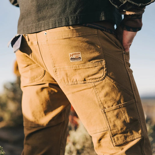 Back view of the Howler Brothers Men's HB Trade Pants in Duck Brown showing the back pocket logo and cargo pocket against a desert background.