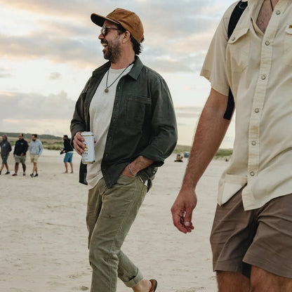 A man wearing the Anthracite Howler Brothers Mens HB Trade Shirt Long Sleeve walks on a beach at sunset with other people in the background.