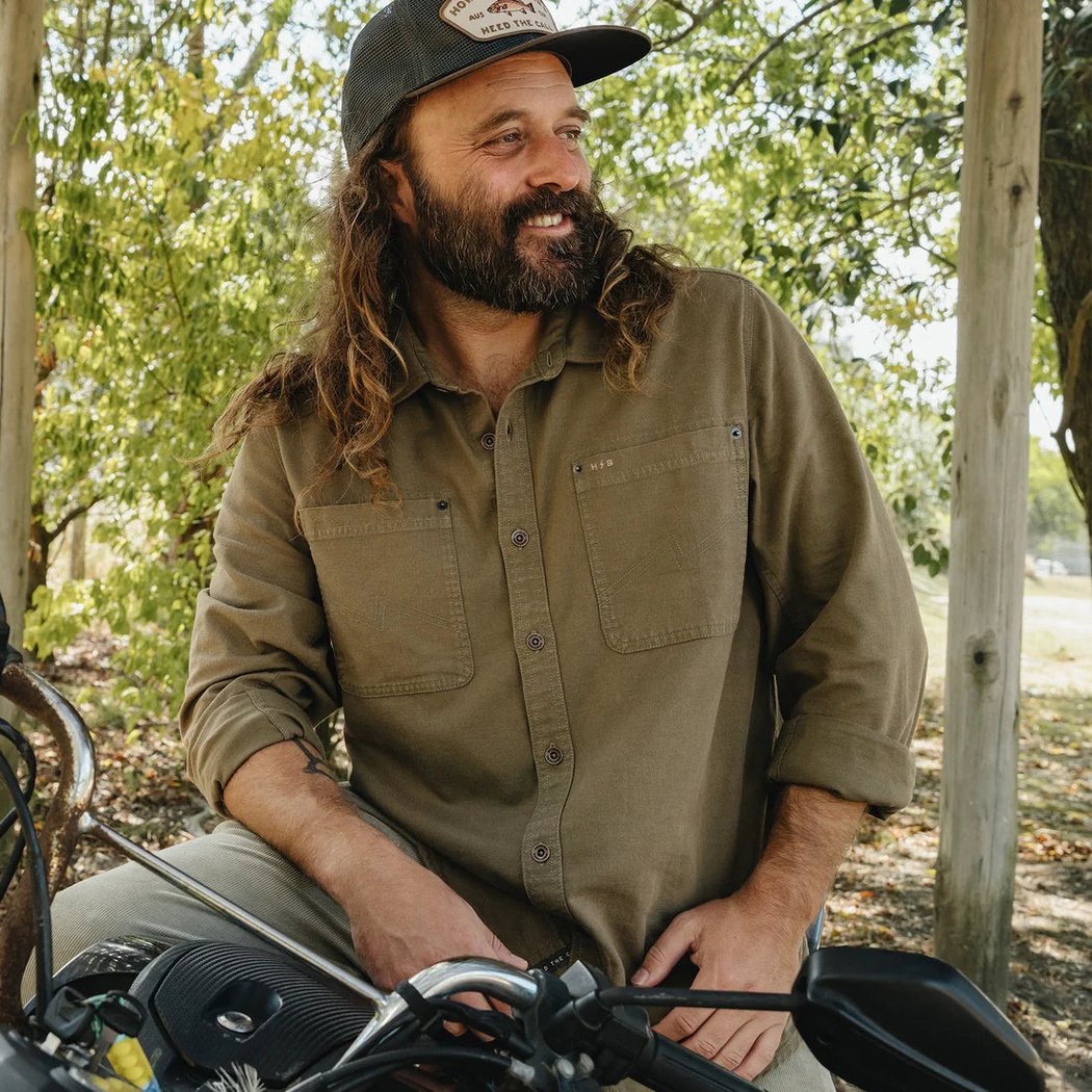 A man wearing the Howler Brothers Mens HB Trade Shirt Long Sleeve in Anthracite sits on a motorcycle in a wooded outdoor setting.