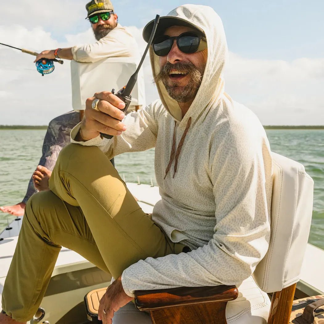 A man wearing the Howler Brothers Men's Loggerhead Hoodie in Meridians Plains sits on a boat with another man fishing in the background.