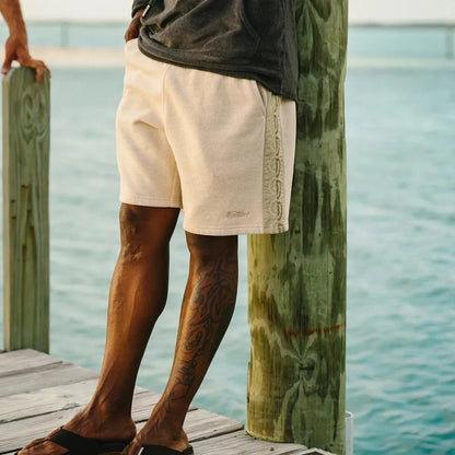 A man wearing Howler Brothers Men's Palmetto Chill Shorts in Oatmeal Heather stands on a dock by the water.
