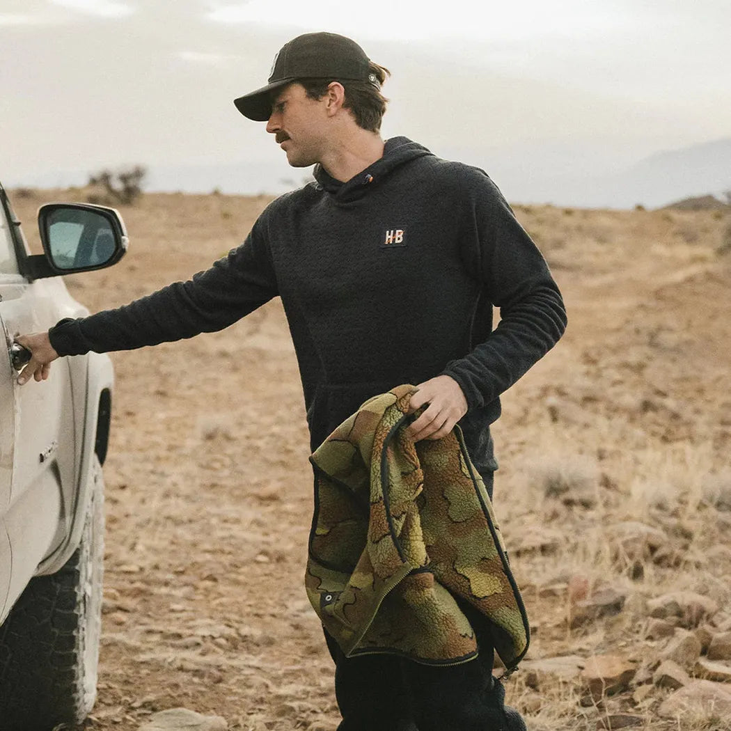 A man in a Howler Brothers Men's Palo Duro Fleece Hoodie stands by a white truck in a rocky, desert-like landscape, holding a camouflage-patterned item.