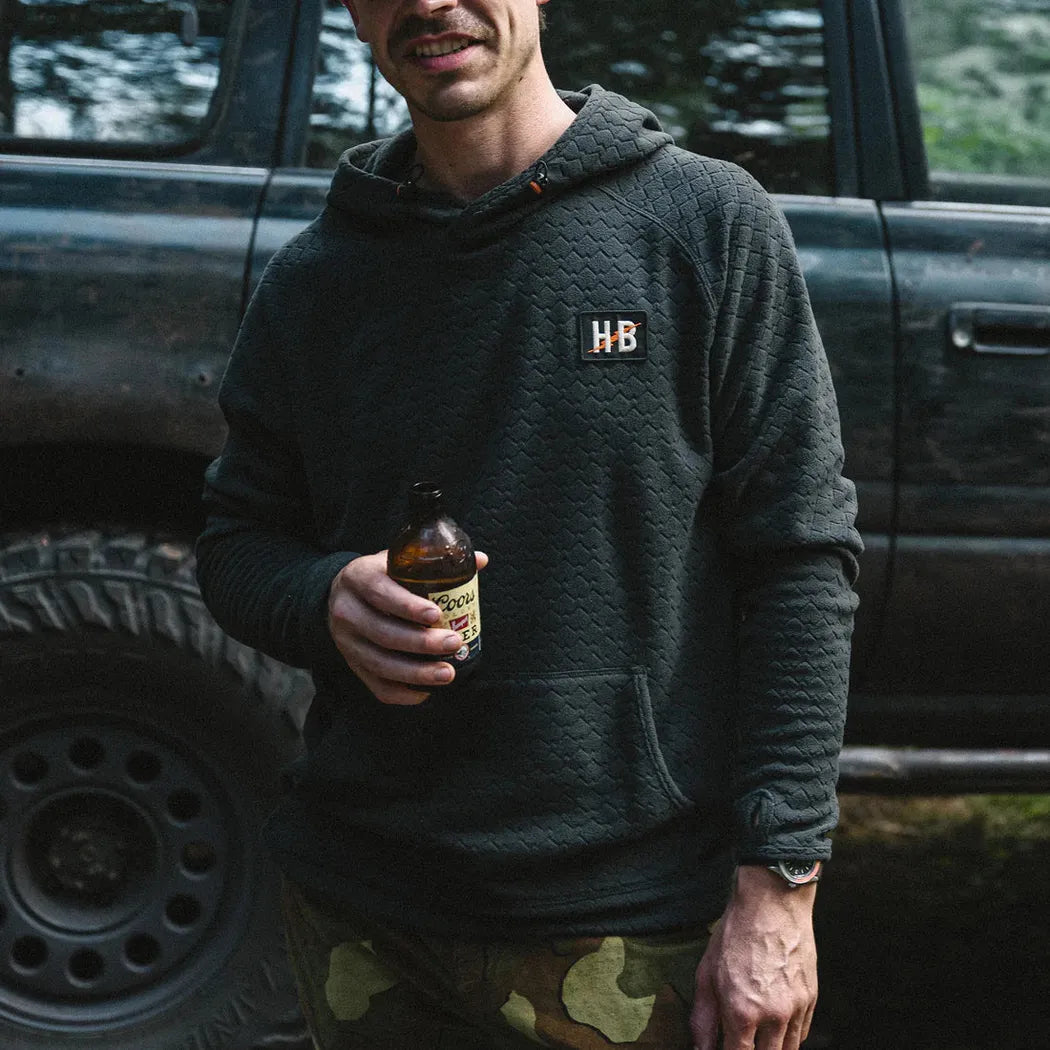 A man in a Mountain Grid : Antique Black Howler Brothers Men's Palo Duro Fleece Hoodie holds a beer bottle in front of a truck.