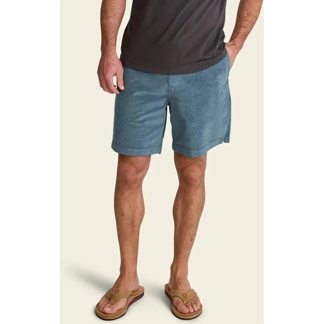 A front view of the Howler Brothers Men's Pressure Drop Cord Shorts in Grid : Blue Mirage, showing a model wearing them with a dark t-shirt and tan flip-flops against a light background.