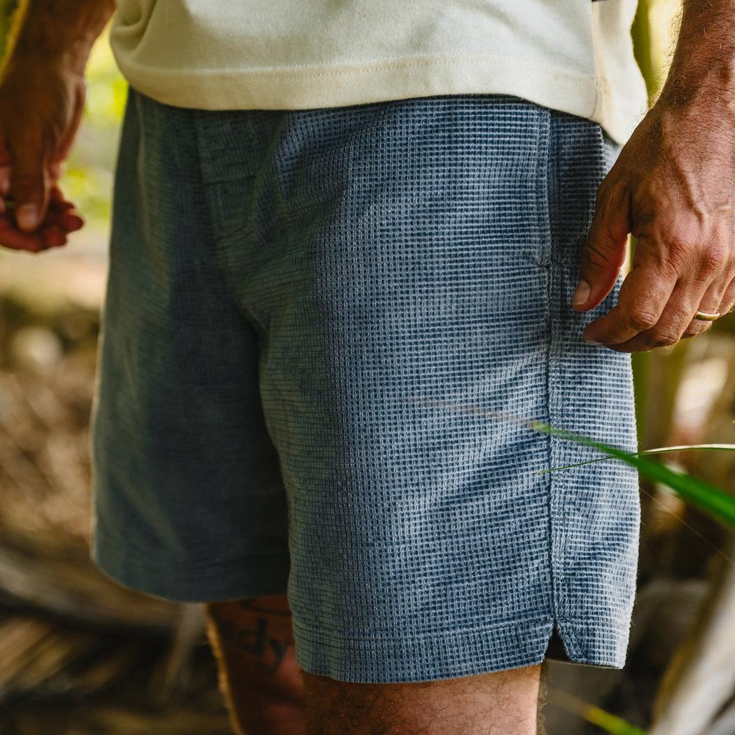 Side view of the Howler Brothers Men's Pressure Drop Cord Shorts in Grid : Blue Mirage showing the side seam and a model wearing them against a natural outdoor background.