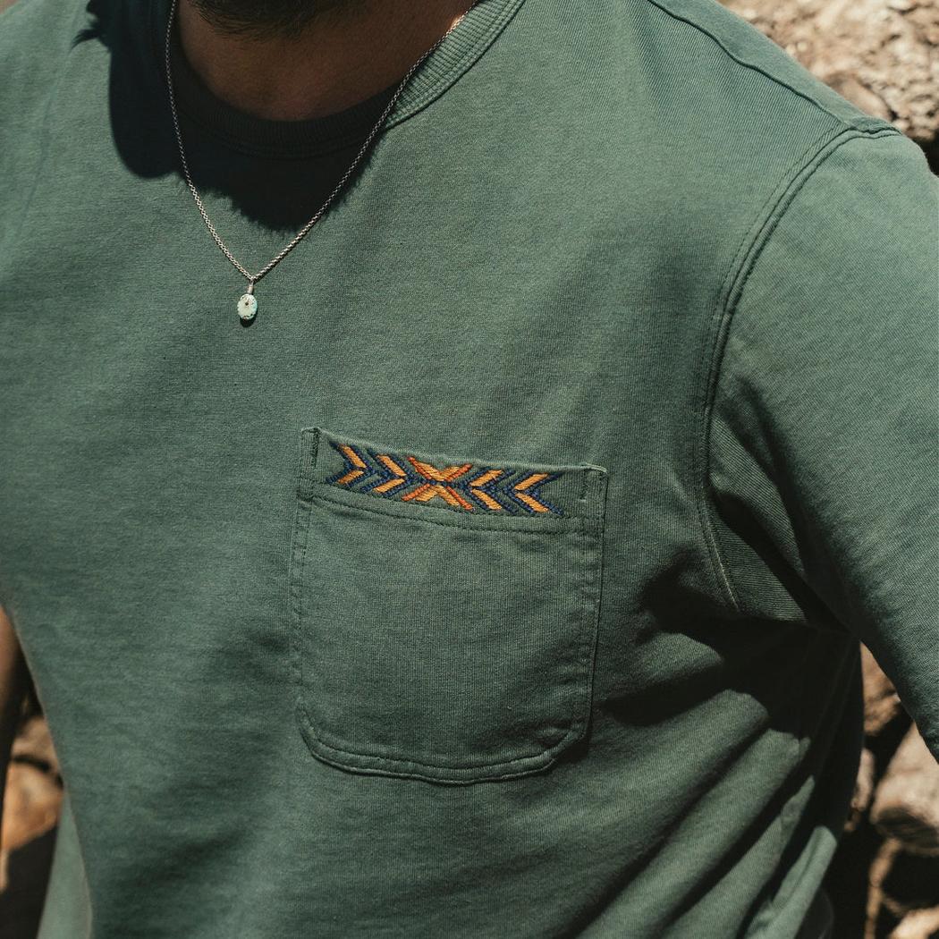 A close-up of the Howler Brothers Men's Spectrum Pocket T in Bone White, showing the pocket embroidery and a model wearing it against a natural background.