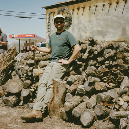 A man in Bone White Howler Brothers Men's Spectrum Pocket T leans on a log with a stack of firewood behind him.