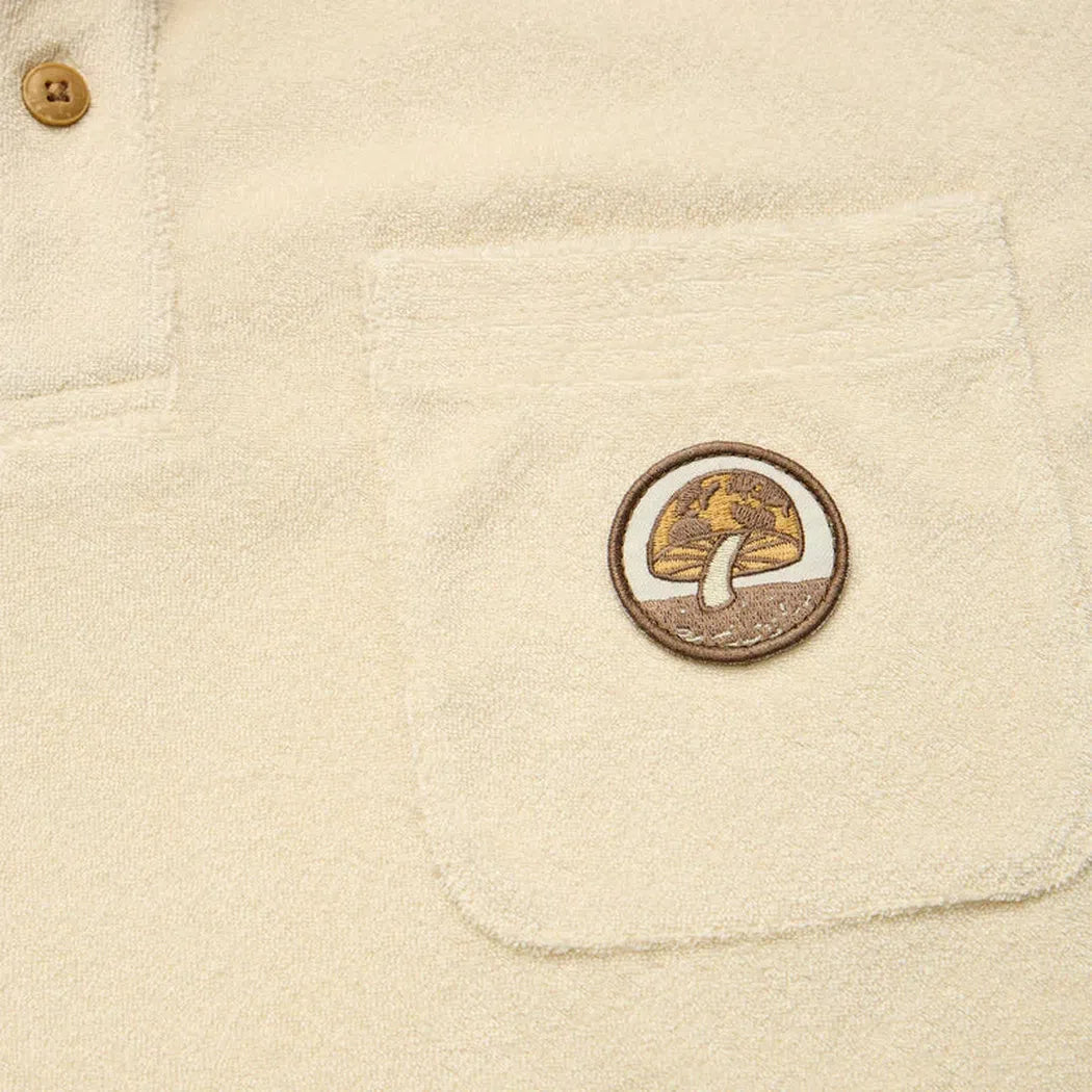 A close-up of the Howler Brothers Plusherman Terry Polo in Cream showing the pocket and patch against a neutral background.