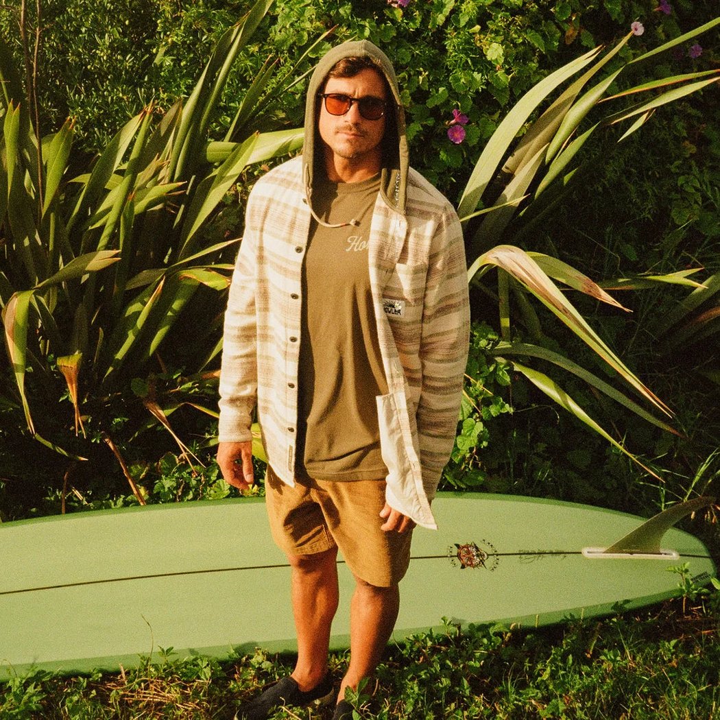 A man wearing the Howler Brothers Salvo Hooded Blanket Flannel in Pathway Stripe Dawn stands outdoors with a surfboard and lush greenery behind him.
