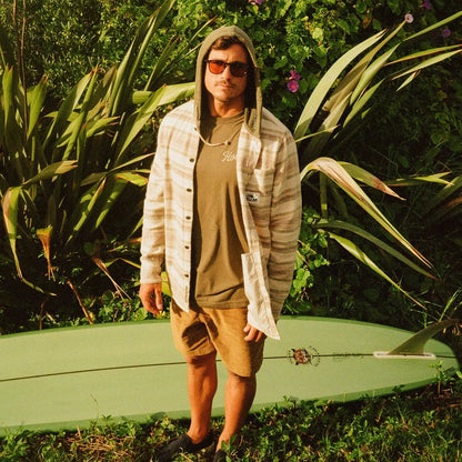 A man wearing the Howler Brothers Salvo Hooded Blanket Flannel in Pathway Stripe Dawn stands outdoors with a surfboard and lush greenery behind him.
