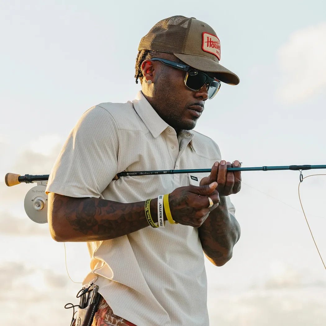 A man wearing a Howler Brothers Standard Hat in Howler Electric Fatigue color holds a fishing rod with a clear sky background.