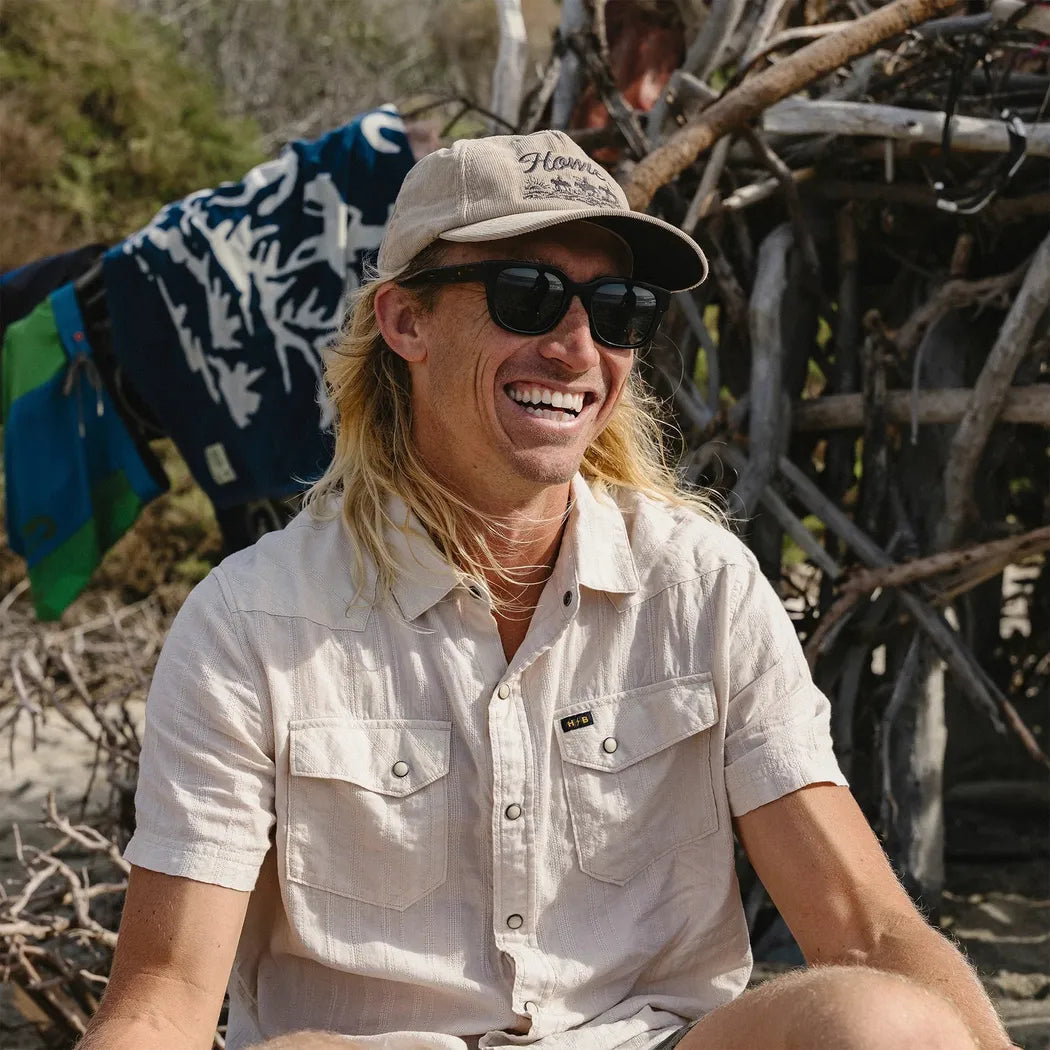 A smiling man with long blonde hair wears the Howler Brothers Standard Hat in Khaki Corduroy, sitting outdoors near driftwood and a blue-and-white patterned blanket.