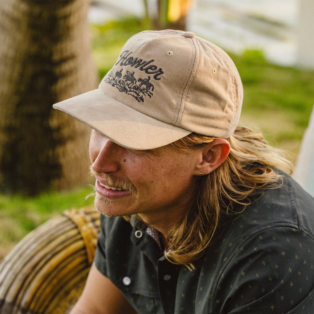 A man with long hair wears the Howler Brothers Standard Hat in Khaki Corduroy, sitting outdoors near a tree.