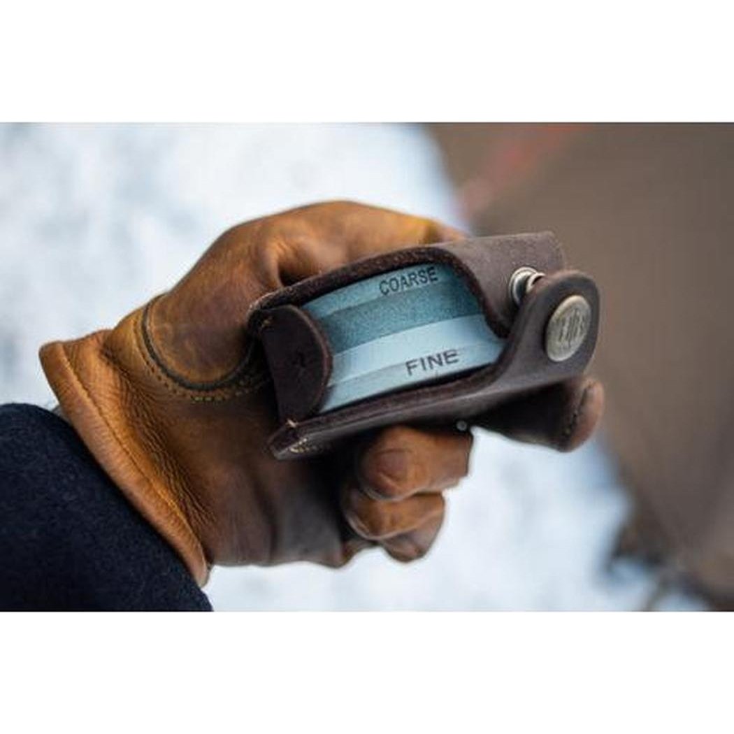 A hand holds the Hults Bruk Grinding Stone 180/600 with "Coarse" and "Fine" visible, against a snowy background.