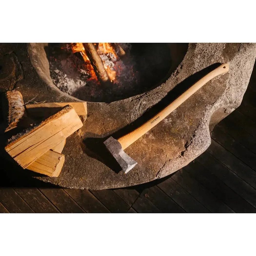 A natural stone fire pit with burning wood, a stack of firewood, and the Hults Bruk Kvillinge Splitting Axe resting on the edge.