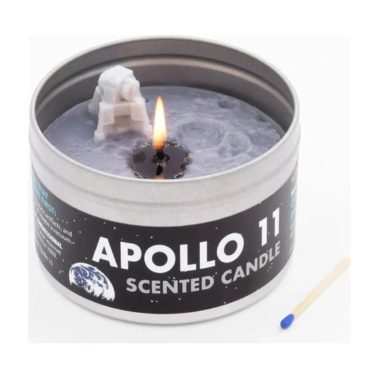 A close-up of the JD and Kate Industries Apollo 11 Scented Candle in a tin with a lit wick and a small astronaut figure, next to a matchstick.