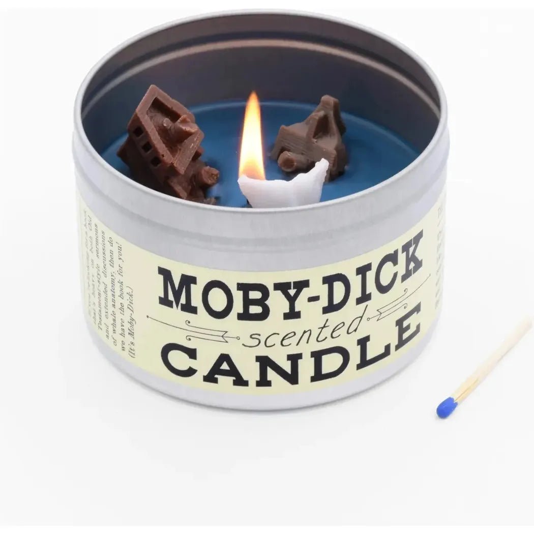A close-up of a JD and Kate Industries Moby Dick Scented Candle in a tin with a lit wick and chocolate house decorations inside, next to a matchstick on a white background.
