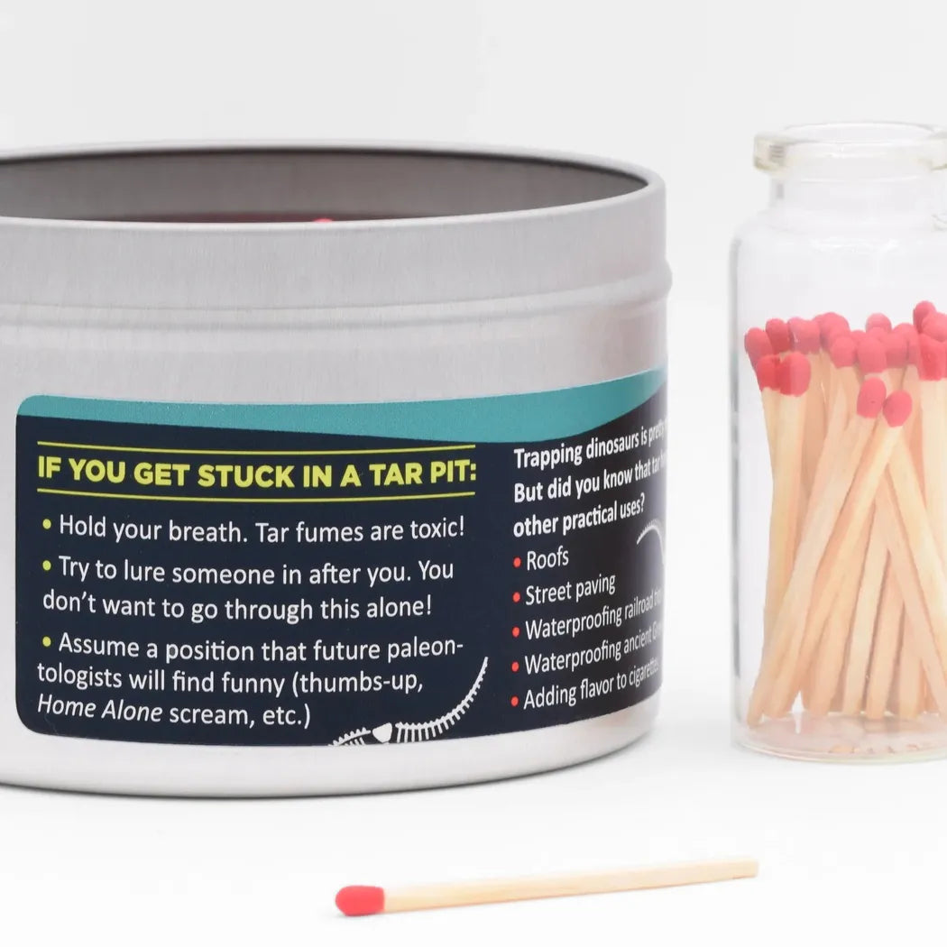 A silver JD and Kate Industries Tar Pit Scented Candle tin with a humorous label sits next to a glass jar of red-tipped matches on a white background.