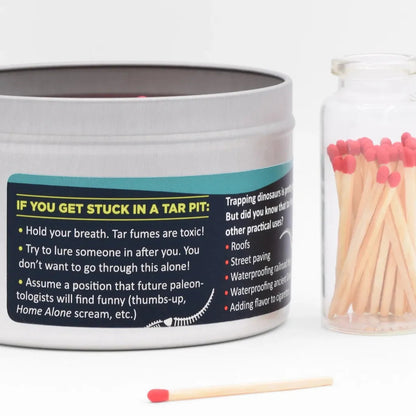 A silver JD and Kate Industries Tar Pit Scented Candle tin with a humorous label sits next to a glass jar of red-tipped matches on a white background.