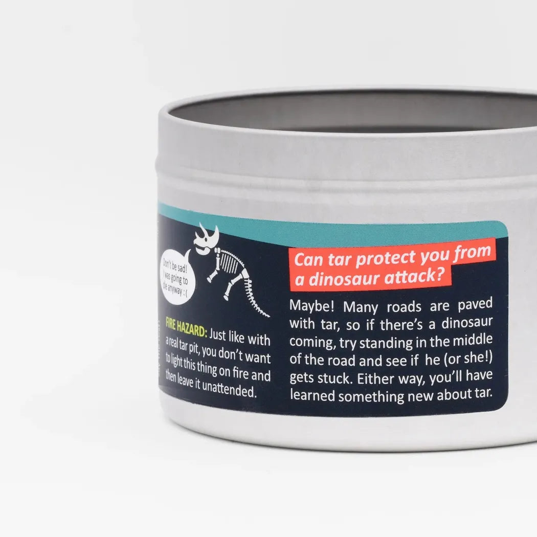 A close-up of the JD and Kate Industries Tar Pit Scented Candle showing its label with a dinosaur graphic and text about tar protection against a white background.