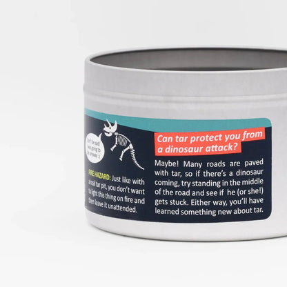 A close-up of the JD and Kate Industries Tar Pit Scented Candle showing its label with a dinosaur graphic and text about tar protection against a white background.