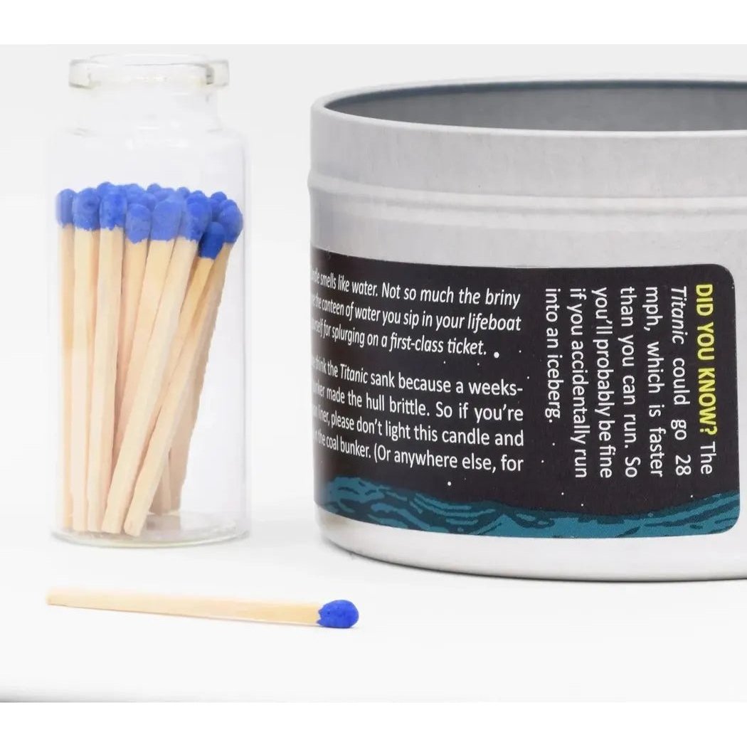 A clear jar of blue-tipped matches sits next to a silver tin of the Titanic scented candle, with one match on the white surface.